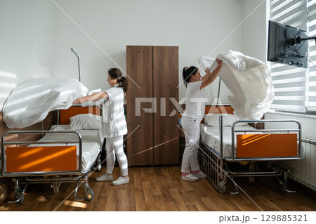 Two nurses in uniforms cleaning and organizing a hospital room by changing the bed linens and ensuring a hygienic environment for patients. 129885321