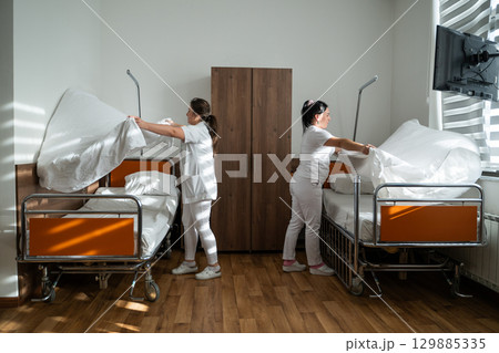 Two nurses in uniforms cleaning and organizing a hospital room by changing the bed linens and ensuring a hygienic environment for patients. Two nurses in uniforms cleaning and organizing a hospital room by changing the bed linens and ensuring a hygienic environment for patients. 129885335