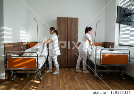Two nurses in uniforms cleaning and organizing a hospital room by changing the bed linens and ensuring a hygienic environment for patients. 129885337