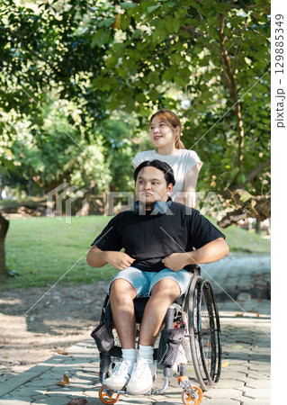 Young Woman Assisting Man in Wheelchair Enjoying a Peaceful Stroll in a Lush Green Park on a Sunny Day 129885349