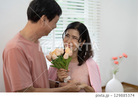 Happy Senior Couple Celebrating Valentine's Day with Flowers and Smiles in Cozy Home Setting Happy Senior Couple Celebrating Valentine's Day with Flowers and Smiles in Cozy Home Setting 129885374