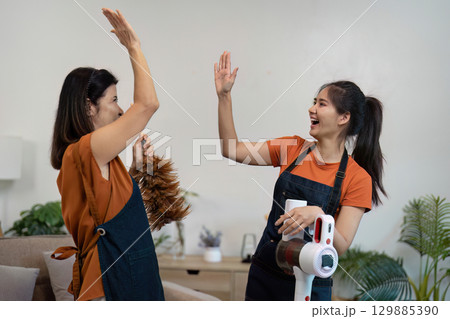 Joyful Women Engaged in House Cleaning with Feather Duster and Vacuum in Modern Living Room Setting 129885390