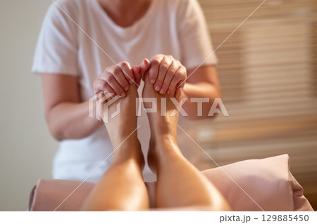 Foot Massage Therapy in a Relaxing Spa Setting. A professional massage therapist in a white uniform provides a soothing foot massage to a client in a calm and serene spa environment 129885450