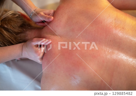 Relaxing Back Massage in a Spa Setting. A professional massage therapist in a blue uniform provides a relaxing back massage to a client lying on a massage table. 129885482