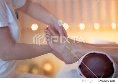 Foot Massage Therapy in a Relaxing Spa Setting. A professional massage therapist in a white uniform provides a soothing foot massage to a client in a calm and serene spa environment 129885486