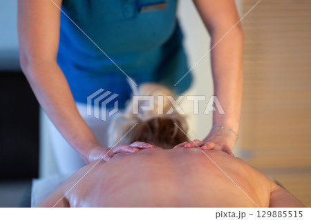 Relaxing Back Massage in a Spa Setting. A professional massage therapist in a blue uniform provides a relaxing back massage to a client lying on a massage table. 129885515