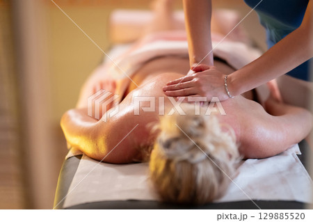 Relaxing Back Massage in a Spa Setting. A professional massage therapist in a blue uniform provides a relaxing back massage to a client lying on a massage table. 129885520