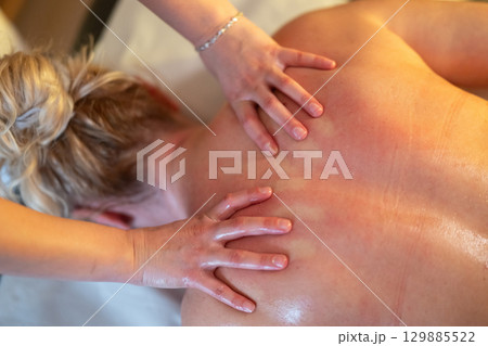 Relaxing Back Massage in a Spa Setting. A professional massage therapist in a blue uniform provides a relaxing back massage to a client lying on a massage table. 129885522