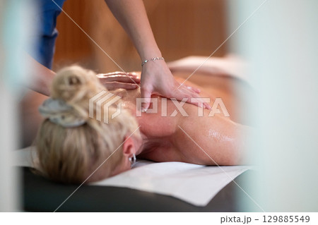 Relaxing Back Massage in a Spa Setting. A professional massage therapist in a blue uniform provides a relaxing back massage to a client lying on a massage table. 129885549