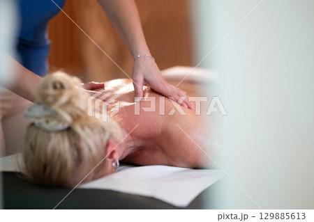 Relaxing Back Massage in a Spa Setting. A professional massage therapist in a blue uniform provides a relaxing back massage to a client lying on a massage table. 129885613