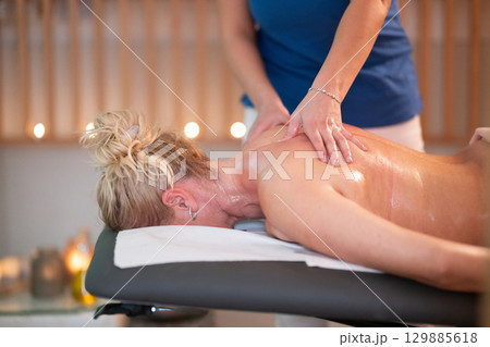 Relaxing Back Massage in a Spa Setting. A professional massage therapist in a blue uniform provides a relaxing back massage to a client lying on a massage table. 129885618