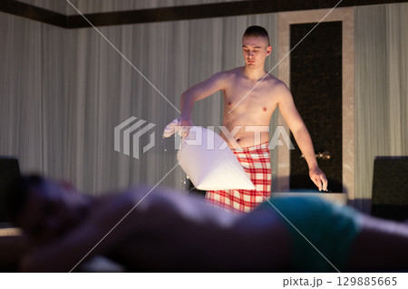 Hamam Attendant with Towel.Young male attendant preparing towel in traditional Turkish bath, standing near client. Hamam Attendant with Towel.Young male attendant preparing towel in traditional Turkish bath, standing near client. 129885665