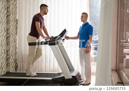 Physical Therapy on Treadmill.Man undergoing physical therapy with trainer assistance on a treadmill indoors. Physical Therapy on Treadmill.Man undergoing physical therapy with trainer assistance on a treadmill indoors. 129885735