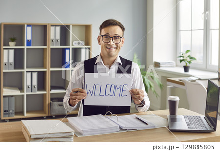 Happy businessman sitting at the desk in office and looking at camera with a message word welcome 129885805