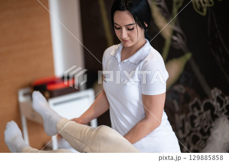 Physiotherapy Session with Blue Foam Roller.A therapist guiding a patient through leg exercises with the help of a blue foam roller in a therapy clinic. 129885858