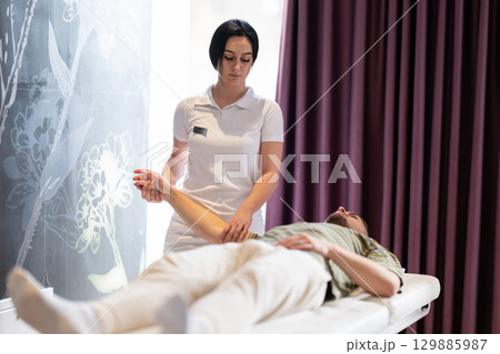 A female therapist performs a arm stretch technique on a male patient lying on a treatment bed. A female therapist performs a arm stretch technique on a male patient lying on a treatment bed. 129885987
