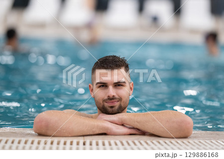 Man resting on the edge of a large indoor pool with a calm expression and relaxed posture. Man resting on the edge of a large indoor pool with a calm expression and relaxed posture. 129886168