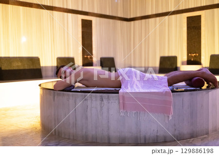 Man relaxing on a marble platform inside a Turkish bath with warm lighting. 129886198
