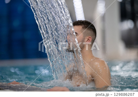 Man standing under a hydrotherapy waterfall in a spa pool, receiving water massage on his head and shoulders. 129886207