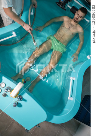Therapeutic Water Bath.Man relaxing in hydrotherapy bath while therapist adjusts water settings. 129886582