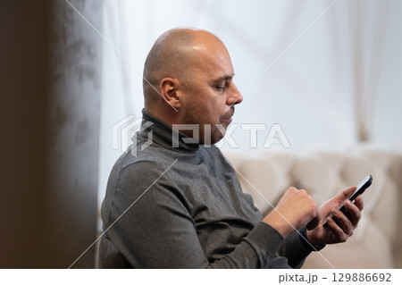 Man Using Smartphone Indoors.Bald man wearing a grey turtleneck using his smartphone while seated indoors. 129886692