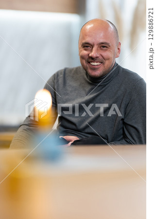 Smiling Man Sitting at Table.Smiling bald man in a turtleneck sweater sitting at a table with a candle in foreground. 129886721