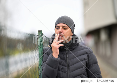 Man Smoking Outside.Man in winter jacket and beanie smoking a cigarette near a chain link fence outdoors. 129886730