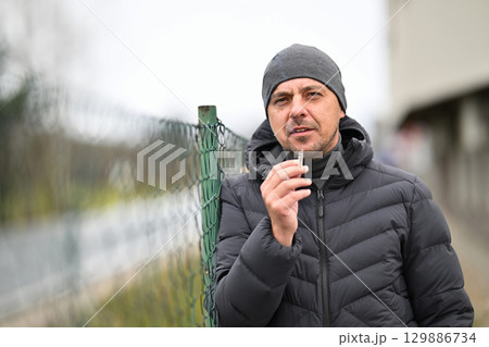 Man Smoking Outside.Man in winter jacket and beanie smoking a cigarette near a chain link fence outdoors. Man Smoking Outside.Man in winter jacket and beanie smoking a cigarette near a chain link fence outdoors. 129886734