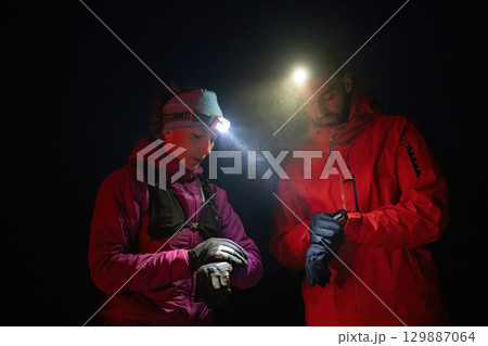 Checking Coordinates During Night Hike.Couple night hikers examine a device under headlamp light, emphasizing navigation and teamwork under low light conditions. 129887064