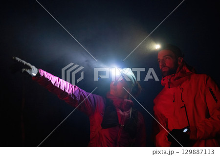 A woman points into the distance while discussing directions with her hiking partner under the beam of headlamps. 129887113