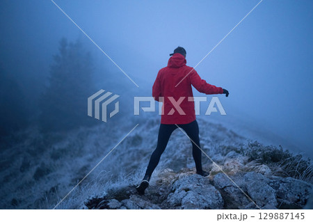 Balancing on the Mountain Trail at Dusk.A lone hiker balances on a rocky mountain trail during early morning or dusk, with a headlamp lighting the misty path. 129887145