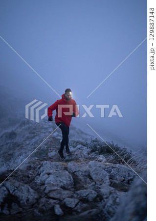Balancing on the Mountain Trail at Dusk.A lone hiker balances on a rocky mountain trail during early morning or dusk, with a headlamp lighting the misty path. 129887148