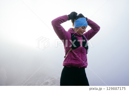 Active female hiker in pink jacket stretching arms on a foggy mountain peak during morning light. 129887175