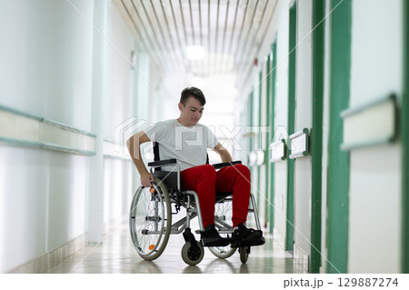 Young teenage boy smiling while sitting in a wheelchair in a hospital hallway. Rehabilitation and positivity concept. Young teenage boy smiling while sitting in a wheelchair in a hospital hallway. Rehabilitation and positivity concept. 129887274