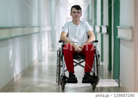 Young teenage boy smiling while sitting in a wheelchair in a hospital hallway. Rehabilitation and positivity concept. 129887280
