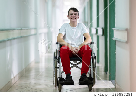 Young teenage boy smiling while sitting in a wheelchair in a hospital hallway. Rehabilitation and positivity concept. 129887281