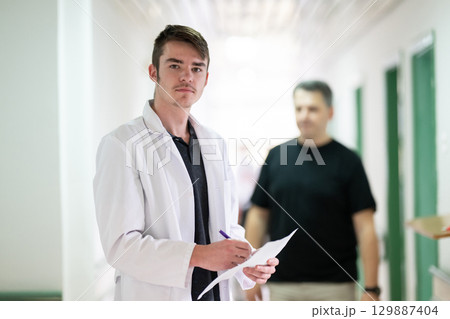 Doctor Reading Documents in Hospital.Young male doctor walking in hospital hallway while reading patient documentation. Clinical work environment. 129887404