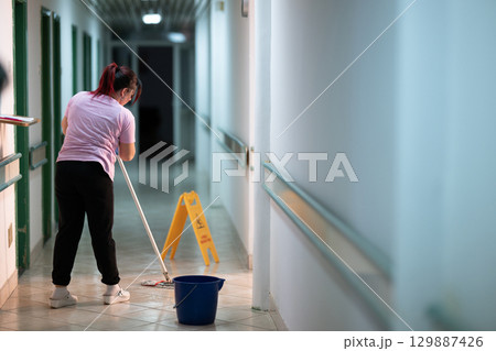 Cleaning Staff in Hospital Hallway.Female janitor standing in a hospital corridor with mop and bucket, symbolizing cleanliness and hygiene. Cleaning Staff in Hospital Hallway.Female janitor standing in a hospital corridor with mop and bucket, symbolizing cleanliness and hygiene. 129887426