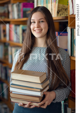 Portrait of a female student with a stack of books in library. Cheerful caucasian girl studying for college exams, prepares class assignment Portrait of a female student with a stack of books in library. Cheerful caucasian girl studying for college exams, prepares class assignment 129888337