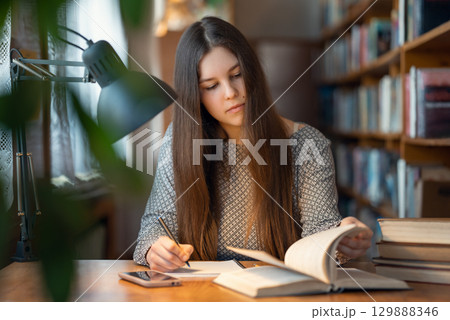 Female student working in library. Focused student learning, getting ready for important seminar, doing home assignment, writing information from book Female student working in library. Focused student learning, getting ready for important seminar, doing home assignment, writing information from book 129888346