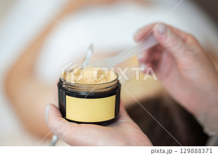 Brown half transparent plastic container with facial mask in female hands, copy space. Professional cosmetologist taking mask to apply in it on clients face 129888371