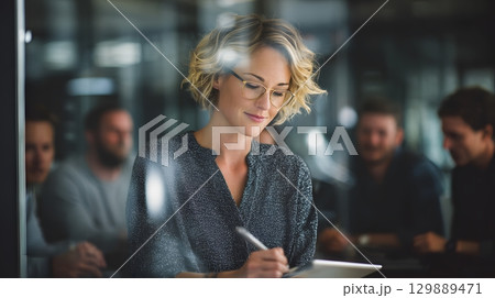 businesswoman writing digital notes on tablet during team meeting in glass office 129889471
