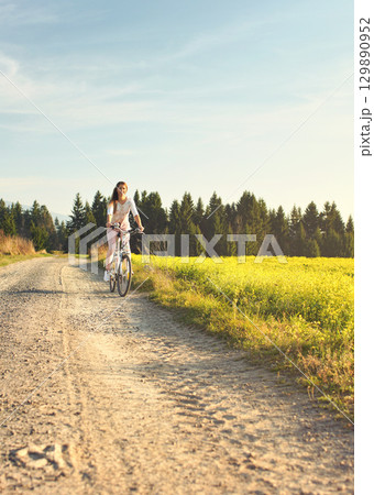 Young woman in summer light clothes rides bicycle on dusty road towards camera, afternoon sun shines to fields and forest in background 129890952