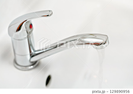 Chrome tap faucet with water flowing, blurred bathroom background 129890956