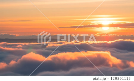 Peaceful sunrise or sunset above fluffy clouds glowing with warm hues and golden sunlight in scenic composition 129891115