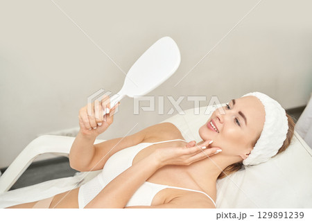 Young caucasian female in a spa chair admiring skin in mirror during relaxing Young caucasian female in a spa chair admiring skin in mirror during relaxing 129891239