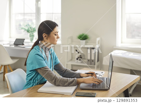 Nurse, young female doctor using laptop online, writing notes in medical journal sitting at desk 129891353