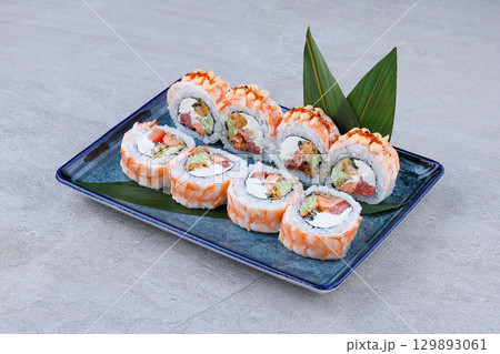 Colorful shrimp-wrapped sushi rolls filled with avocado, cream cheese, and assorted seafood, topped with spicy mayo and served on a blue plate. 129893061
