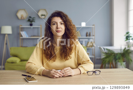 Young curly woman sitting at home at the desk looking at the camera having online video call. Young curly woman sitting at home at the desk looking at the camera having online video call. 129893849