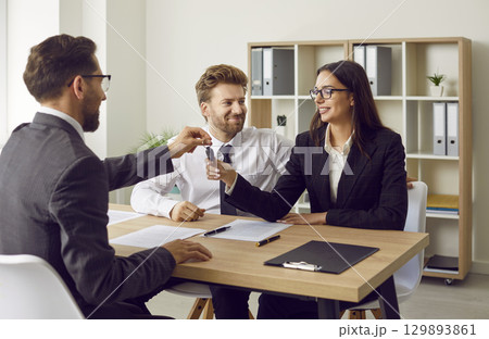 Man realtor giving house or car key to a couple customers sitting in the office. Man realtor giving house or car key to a couple customers sitting in the office. 129893861